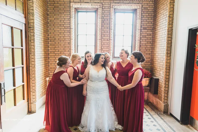 A bride with five bridesmaids in deep red dresses beside the tall arched windows